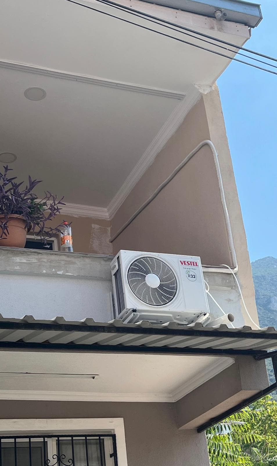 Balkon parapetine konsollanmış Vestel Inverter R32 dış ünite; arkada açık gökyüzü ve dağ manzarası